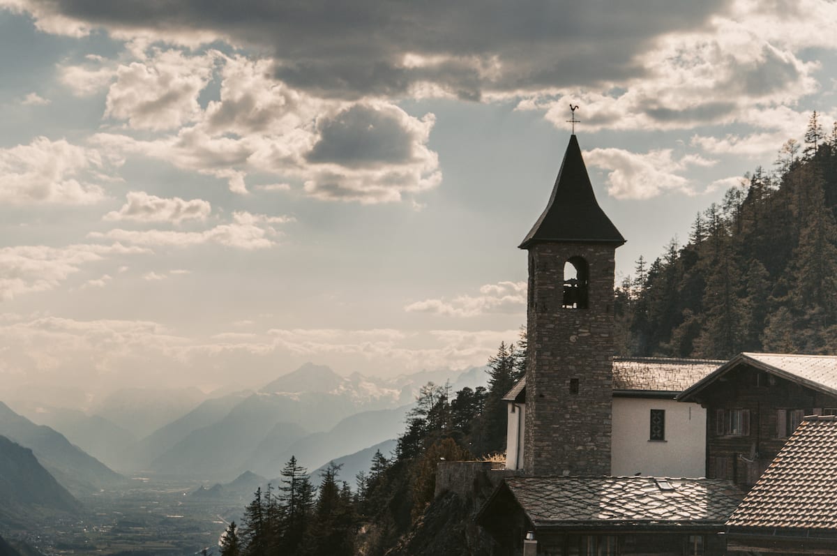 Kirchenturm in Guttet im Wallis. Im Hintergrund ist eine umrisshafte Bergkulisse zu erkennen.