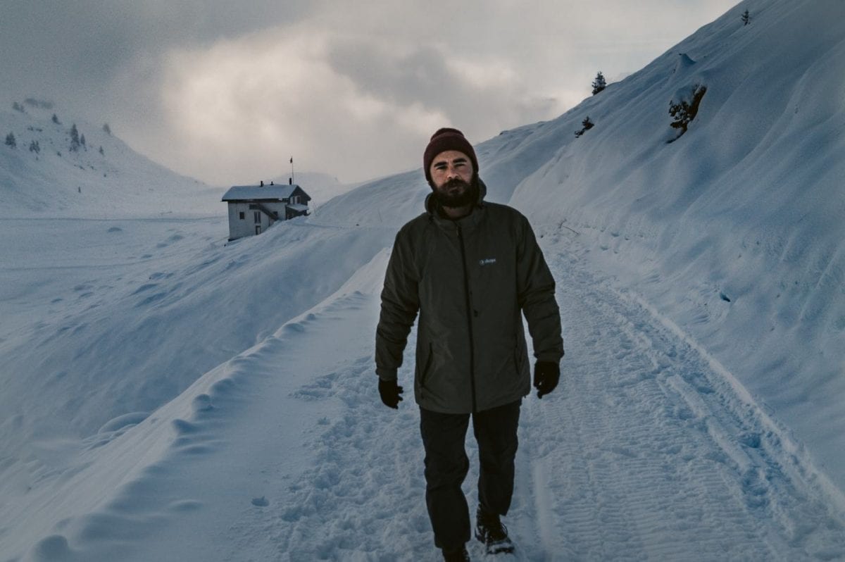 Bertal geht durch den Schnee auf einem Winterwanderweg in der Schweiz. Hinter ihm ist in der Ferne ein kleines Haus zu sehen.